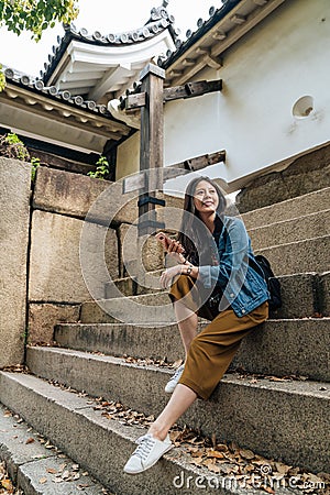 Young happy girl on backpacking vacation sitting onÂ the stairs in Japan. elegant traveler waiting for the tour guide in osaka Stock Photo